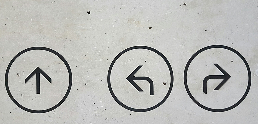 Auf einer Betonwand sind drei Pfeilsymbole zu sehen. Diese stehen jeweils in einem Kreis und zeigen geradeaus und nach links und rechts.