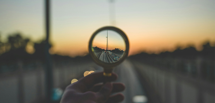 Eine Hand hält eine Lupe, die auf eine Straße im Sonnenuntergang gerichtet ist. Während das Bild im Ganzen unscharf ist, ist der Bereich in der Lupe scharf.