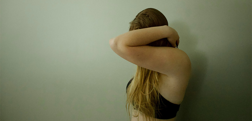 Eine junge Frau mit langen blonden Haaren steht in der Seitenansicht mit einem Bustier bekleidet. Das Gesicht ist nicht zu sehen, da sie den Arm angewinkelt vor selbiges hält.