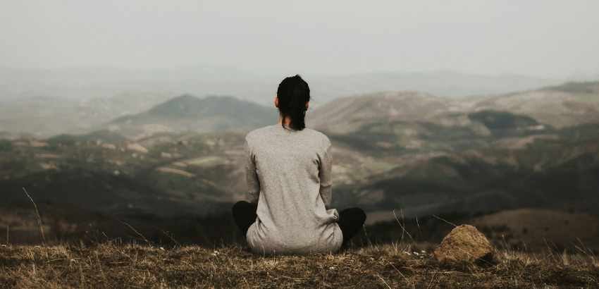 Eine Frau sitzt im Schneidersitz auf dem Boden in einer bergigen Landschaft und schaut in die Ferne. 