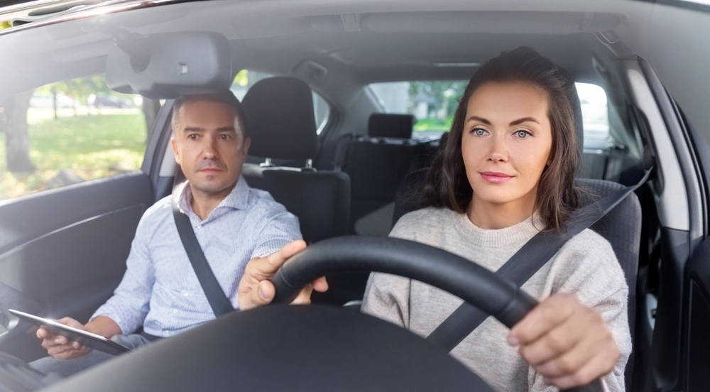 Eine Frau sitzt am Steuer eines Autos. Neben ihr sitzt ein Mann mit Tablet in der Hand, der korrigierend ans Lenkrad fasst.